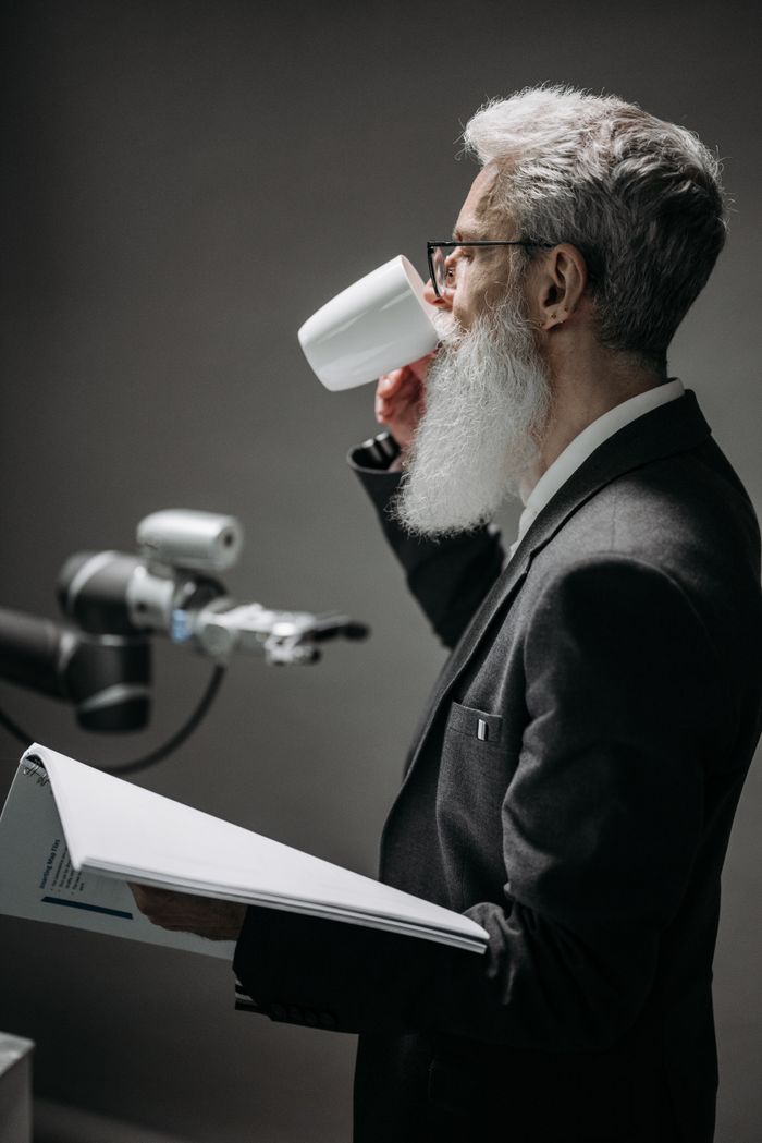 A researcher in formal attire sips coffee beside a robotic arm in a cutting-edge laboratory.